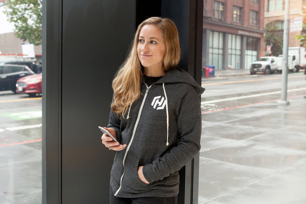 A woman wearing a dark gray zip-up hooded sweatshirt with drawstrings and the white HashiCorp H icon on her left chest.